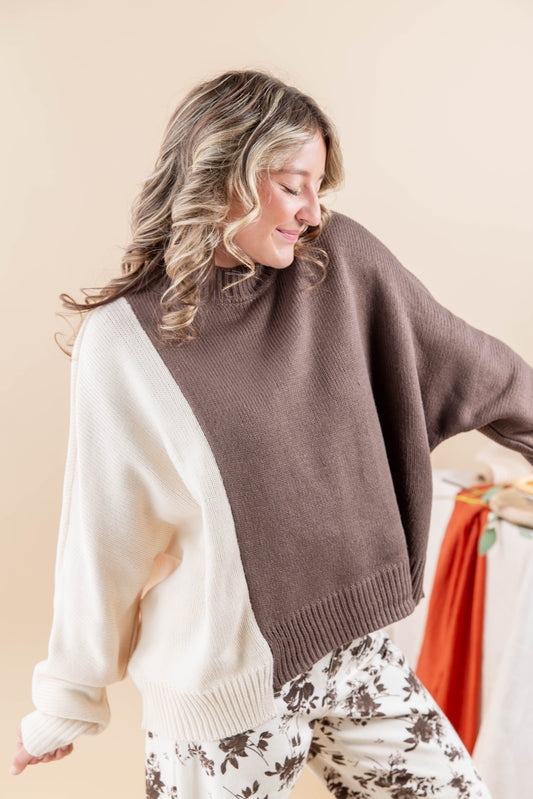 A woman wearing a color-block knit sweater with a taupe brown & cream sleeves, paired with cream and brown floral print pants, set against a warm beige background.
