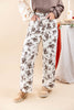 A woman wearing wide-leg white pants with a dark brown floral pattern, styled with a beige sweater and tan loafers, standing against a warm beige background.