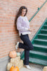 A woman posing outdoors against a brick wall and green stairs, wearing a light lavender ribbed knit sweater, dark navy pants, and white ankle boots, with pumpkins and hay bales at her feet.