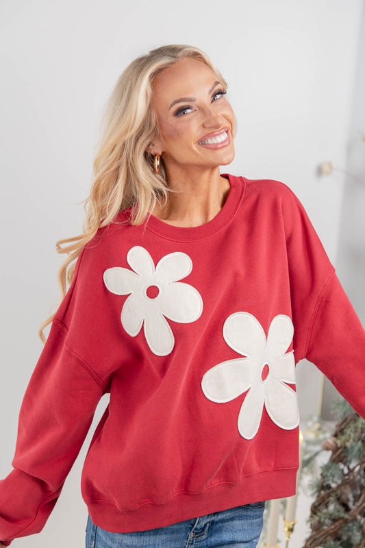 A blonde woman wearing a red crewneck sweatshirt with large white flower appliques, paired with blue jeans.