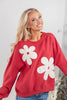 A blonde woman wearing a red crewneck sweatshirt with large white flower appliques, paired with blue jeans.