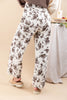 A woman wearing white wide-leg pants with a dark brown floral print, styled with a brown top and beige platform sandals in a warm beige studio setting.