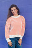 A smiling brunette woman wearing a Halloween inspired look with white knit sweater with orange vertical stripes and denim, standing against a bold purple wall.