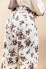 A close-up of a woman wearing high-waisted, wide-leg trousers with a brown floral toile pattern on an off-white background, styled with a cream knit top and a taupe cardigan.