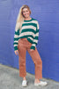 A woman stands against a bright purple brick wall wearing a green and cream striped knit sweater, rust-colored cropped jeans, and white slip-on shoes.