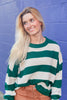 A smiling blonde woman wearing a green and off-white horizontally striped knit sweater, paired with orange pants, standing against a purple painted brick wall.