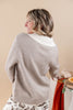 A woman shown from the back wearing a light taupe knit sweater over a white mock neck top, set against a solid beige background.