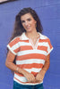 A woman with long dark hair smiling in front of a purple brick wall, wearing a short-sleeve knit polo shirt with horizontal orange and white stripes, a white collar, and a split V-neck.