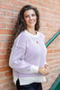 A smiling woman wearing a ivory knit sweater with thin purple vertical stripes and black pants, standing outdoors against a brick wall.