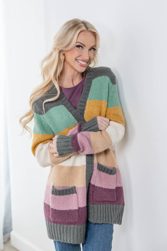 A smiling blonde woman wearing a chunky knit cardigan with large color-blocked stripes in gray, mint green, mustard yellow, cream, pink, and beige, paired with a purple top and blue jeans, standing against a white wall.