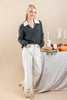 A woman models a dark green v-neck sweater with a contrasting white collar, paired with white wide-leg pants featuring gold floral embroidery at the hem. She is posed next to a styled table with autumn decorations, candles, and a pie, set against a solid beige studio background.