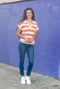 A woman with long dark hair smiling in front of a purple brick wall, wearing a short-sleeve knit polo shirt with horizontal orange and white stripes, a white collar, and a split V-neck.