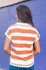 A woman viewed from behind wearing a short-sleeve knit top with bold orange and white horizontal stripes, standing against a blue painted brick wall.