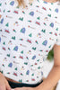 A close-up of a woman wearing a white ribbed knit top with a colorful Christmas-themed print featuring snowman, Santa Claus, snowmobiles, pine trees, and houses.