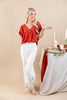 A woman modeling a rust-orange wrap-style blouse and white wide-leg pants with gold floral embroidery near the hem, standing beside a styled table with candles and greenery against a beige studio backdrop.