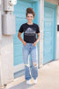 A woman with stands in front of a teal door wearing a black-colored graphic t-shirt with the words 'Sanderson Witch Museum, Home of the Black Flame Candle' printed in white text. She is also wearing blue jeans and white sneakers