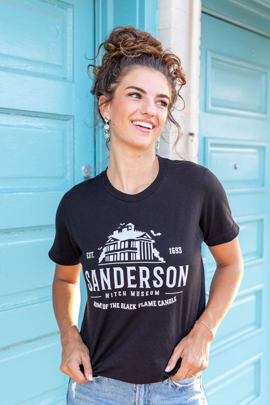 A woman with stands in front of a teal door wearing a black-colored graphic t-shirt with the words 'Sanderson Witch Museum, Home of the Black Flame Candle' printed in white text. She is also wearing blue jeans.