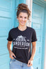 A woman with stands in front of a teal door wearing a black-colored graphic t-shirt with the words 'Sanderson Witch Museum, Home of the Black Flame Candle' printed in white text. She is also wearing blue jeans.