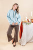 A woman modeling a light blue ribbed knit sweater with a neck tie scarf, paired with brown wide-leg pants and black flats, standing in front of a beige studio backdrop next to a decorated autumn table.