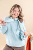A smiling woman with wavy blonde hair wears a light blue ribbed knit sweater with a large bow detail at the neckline, paired with brown pants, against a warm beige studio background.