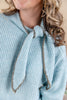 A close-up of a woman wearing a light blue ribbed knit sweater with a knotted neck and textured gray trim along the edges.