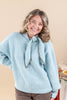 A smiling woman with shoulder-length wavy brown hair wearing a light blue ribbed knit sweater with a tie-neck detail and brown pants, standing against a solid beige background.