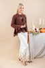 A woman models a brown long-sleeve blouse with a tie-neck and flared ruffle hem, paired with white wide-leg pants featuring gold floral embroidery near the hem. She stands in front of a beige studio backdrop next to a styled autumn table with candles, pumpkins, and a pie.