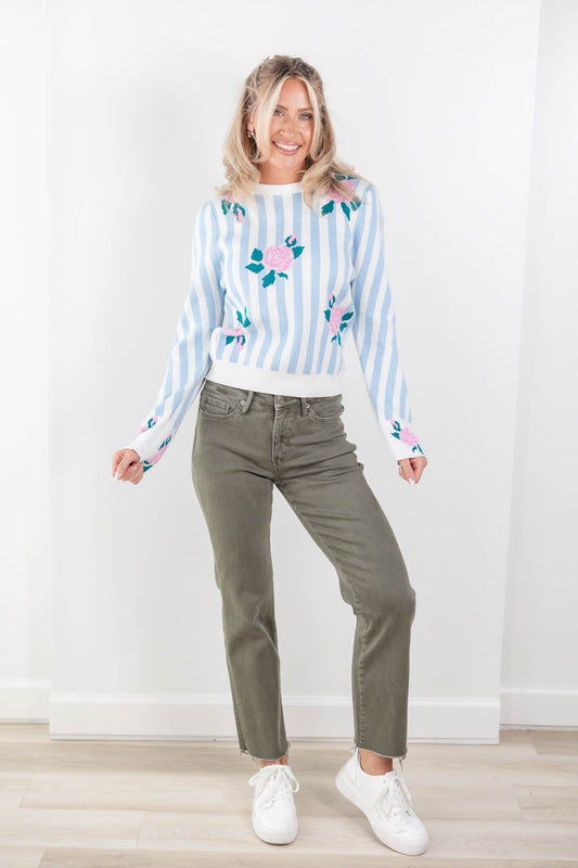 A woman modeling a light blue and white striped crewneck sweater with pink and green floral embroidery, paired with olive green jeans and white sneakers, standing against a white wall.