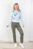 A woman modeling a light blue and white striped crewneck sweater with pink and green floral embroidery, paired with olive green jeans and white sneakers, standing against a white wall.