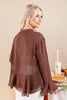 A woman wearing a brown sheer long-sleeve blouse with a ruffled hem, shown from the back in a studio setting with a beige background and a small table with a candle and pie nearby.