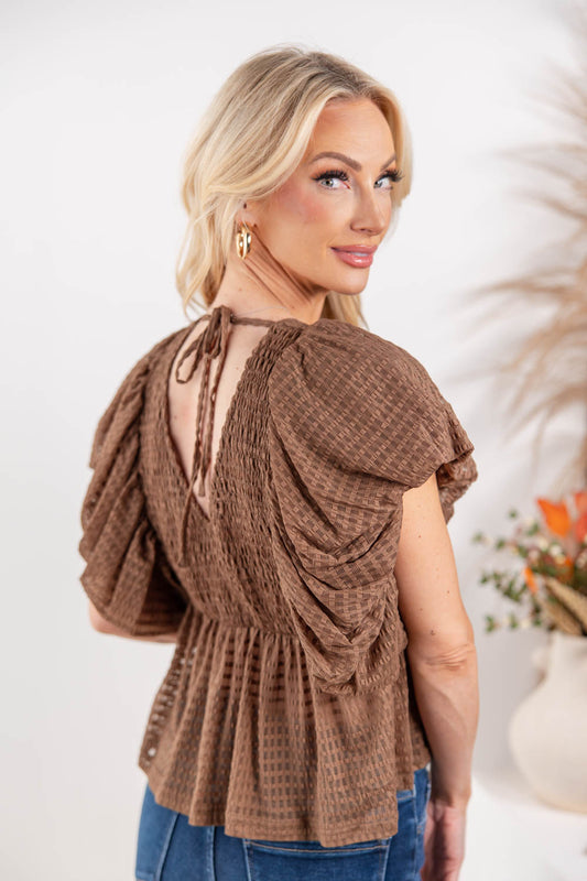 A woman wearing a brown textured short-sleeve top with puff sleeves and a tie-back neckline.