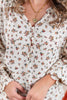 A close-up of a woman wearing a cream-colored blouse with a dense small floral print in orange, green, and red. The blouse features a smocked neckline, a button-down front, and elastic ruffled cuffs.