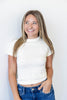 A smiling young woman with blonde-highlighted hair wearing a textured off-white high crew neck short-sleeve crop top and blue jeans, standing against a plain white wall.
