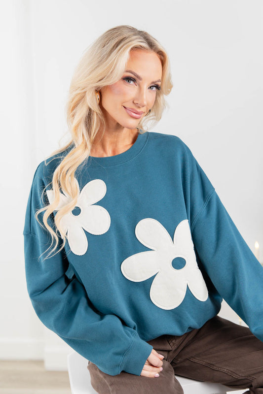 A woman wearing a teal blue sweatshirt with large white flower appliqués.