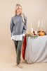 A woman models a gray knit sweater with a contrasting white hem, paired with olive green pants, standing next to a styled autumn table with pumpkins, candles, and a pie.