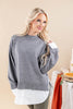 A blonde woman wearing a heather gray knit sweater layered over a white collared shirt, standing against a warm beige studio background with a styled table visible on the right.