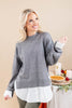 A smiling blonde woman models a gray knit sweater with dramatic wide sleeves and contrasting white cuffs, layered over a white collared shirt. She is posed in front of a warm beige background with a styled table featuring a pie, candles, and autumn decorations.