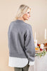 A woman shown from the back wearing a gray knit sweater layered over a white collared shirt, standing in a softly lit indoor setting with candles and a decorated table in the background.
