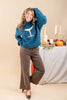 A woman models a teal knit sweater featuring a white martini glass graphic on the chest, paired with brown wide-leg pants and black heels, posed in front of a decorated table with pumpkins and candles.