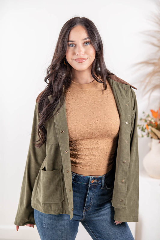 A woman modeling an olive green utility jacket with brown collar, worn over a tan top and blue jeans.