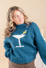 A woman wearing a teal blue knit sweater with a white martini glass graphic and a small colorful olive on the rim, paired with brown pants against a warm beige background.