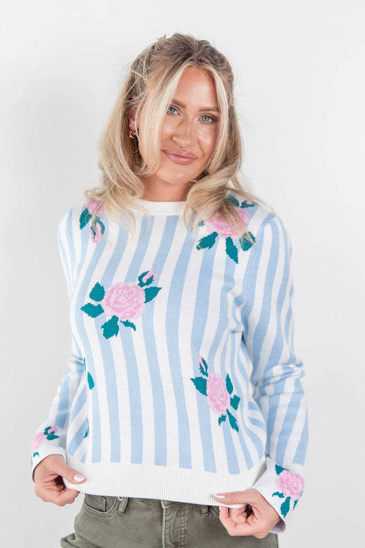 A woman wearing a light blue and white vertically striped sweater with pink roses and green leaves embroidered on the front, paired with olive green pants, against a plain white studio background.