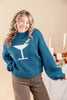 A woman wearing a teal blue knit sweater with a white martini glass graphic and a small yellow oliver on the chest, paired with brown pants, posing in front of a warm beige background with a decorated holiday table.