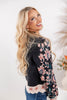 A smiling blonde woman wearing a black knit sweater with pink, blue, and white floral embroidery on the sleeves and a white scalloped hem, paired with blue jeans against a bright white background.