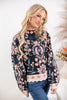 A blonde woman wearing a black crewneck sweater with a colorful floral and paisley pattern in pink, teal, and blue, paired with blue jeans, photographed against a bright white studio background.