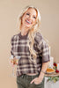 A smiling blonde woman wearing a brown and beige plaid short-sleeve knit top, holding a martini glass in a warm indoor setting.