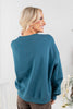 A woman shown from the back wearing a teal blue crewneck sweatshirt and brown.