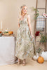 A woman models a sleeveless, floor-length midi dress with a beige and olive green autumn floral pattern, standing in a styled studio setting with pumpkins, a ladder, and a table.