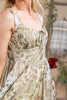 A close-up of a woman wearing a sleeveless midi dress with a beige, sage green, and brown paisley and floral print. The dress features a gathered bodice, thin shoulder straps, and a ruffled tie at the neckline.