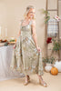 A woman models a sleeveless, midi-length dress with a beige and olive green botanical and stripe pattern, standing in a styled studio setting with autumn props like pumpkins, a ladder, and a table.